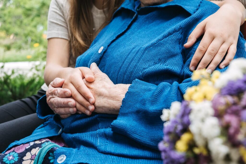 Die gefalteten Hände einer alten Frau werden liebevoll von einer Pflegerin gehalten.